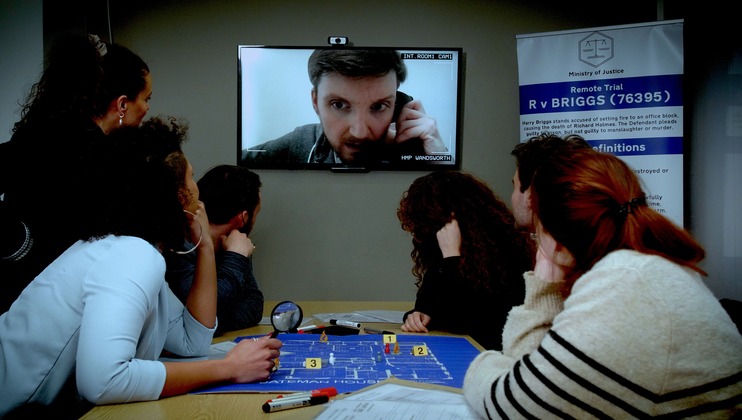 A group of people are gathered around a table, watching a man on a video call displayed on a wall-mounted monitor. The table holds papers and a map with markers. A banner nearby reads Ministry of Justice, mentioning Remote Trial and providing case details.