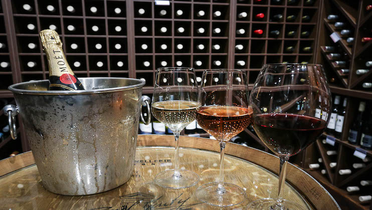 A wine cellar with a metal ice bucket holding a bottle of Moët & Chandon champagne. In front of the bucket are three wine glasses filled with different types of wine: white, rosé, and red. The background shows shelves lined with various wine bottles.