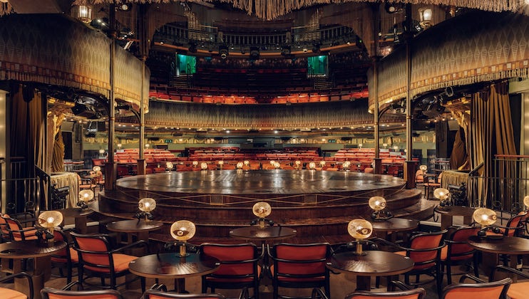 A lavish, empty theater with a round stage and multiple levels of seating. Tables with lamps line the main floor, and curtains, wooden panels, and elaborate decor enhance the opulent ambiance.