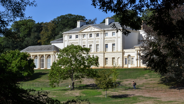 A large, elegant white building with a grand facade stands amidst a lush green landscape. The building features tall windows and intricate architectural details. Trees frame the scene, and a few people are walking and sitting on the lawn in front of the building.