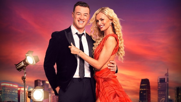 A couple, dressed formally, stand closely together against a vibrant cityscape backdrop at sunset. The man wears a dark suit and smiles warmly, while the woman, in a red dress, rests her hand on his shoulder and smiles. Studio lights and skyscrapers are visible in the background.