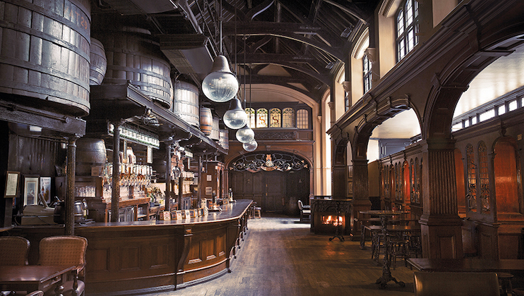 A vintage-style pub interior with wooden barrels, a curved bar counter, high wooden ceilings, and globe-shaped hanging lights. The space features wooden tables and chairs and a mix of warm tones, creating a cozy and inviting atmosphere.
