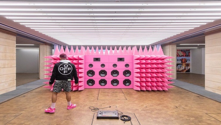 A person wearing a black jacket with a white logo, patterned shorts, pink sneakers, and a pink cap is standing in front of a large, pink, spiked installation resembling speakers. The installation is placed in a well-lit room with a beige-tiled floor.