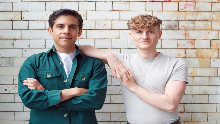 Two people are standing next to each other against a tiled wall. The person on the left, in a green jacket and white shirt, is smiling with crossed arms. The person on the right, in a light gray shirt and necklace, is smiling and resting their hand on the other's shoulder.