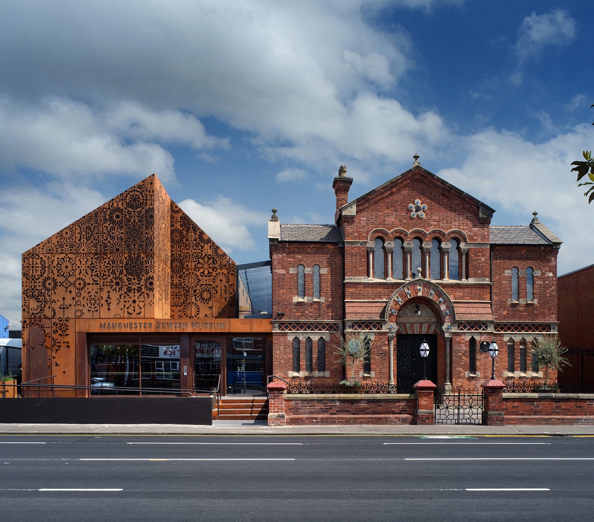 The image shows the Manchester Jewish Museum, featuring a striking blend of historical red-brick architecture and a modern extension with intricate metalwork. The museum's name is displayed prominently, and large windows reveal the building's contemporary design inside.