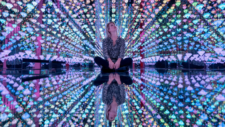 A woman sits in wonder, surrounded by countless bright, multicoloured lights that are reflected by a mirrored floor inside a huge and encompassing immersive art installation in MOCO (The Museum of Modern and Contemporary Art)