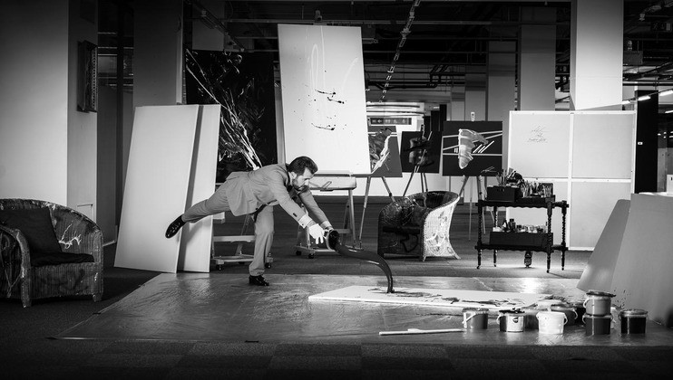 A man in a suit is dramatically pouring paint from a can onto a large canvas on the floor in an art studio. Easels with artworks, paintings, and other art supplies are visible in the background. The scene is captured in black and white.