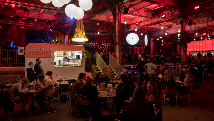 A lively indoor event features a crowd of people sitting at tables, some engaged in conversation. A doughnut truck labeled KRUM Doughnuts is visible on the left. The venue is adorned with colorful lights and hanging decor, with a large clock displaying the time.