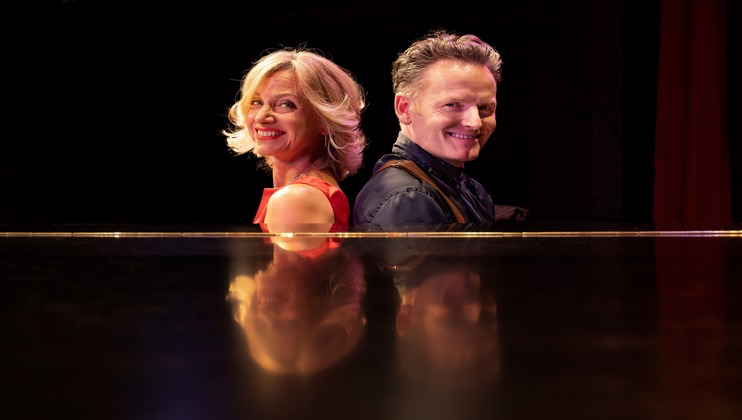 A woman and a man are sitting back-to-back, both smiling towards the camera. The woman has shoulder-length blonde hair and is wearing a red outfit. The man has short, styled hair and is wearing a dark outfit. They are reflected on a glossy surface in front of them.