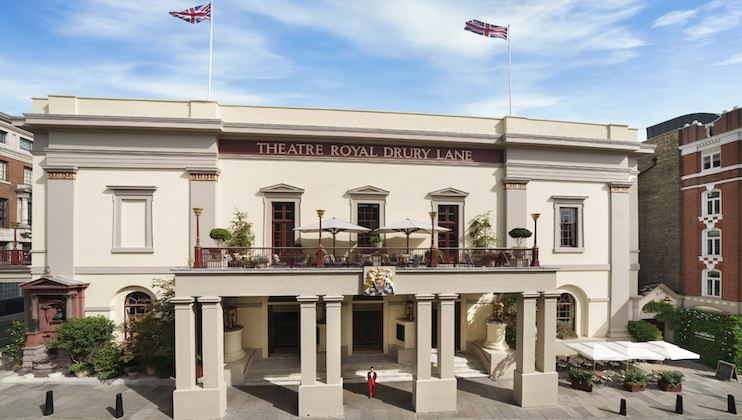 Wide shot of Theatre Royal Drury Lane's façade on a bright day. The neoclassical building features tall columns, two Union Jack flags flying atop, and a balcony with outdoor seating. A person in red stands in front of the grand entrance.