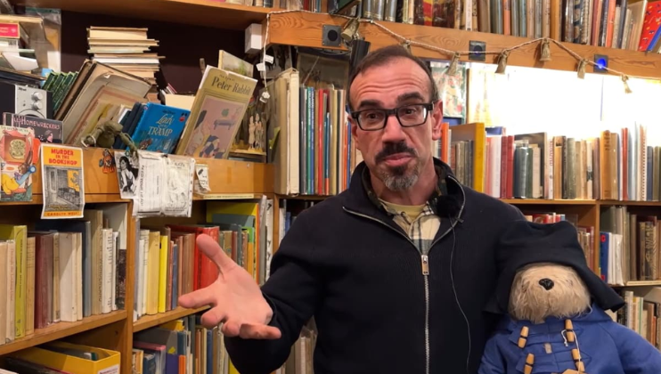 A man with glasses and a beard stands in a cluttered bookstore, holding a Paddington Bear toy in his left hand. He gestures with his right hand while surrounded by shelves filled with books and various items. The lighting is bright and warm.