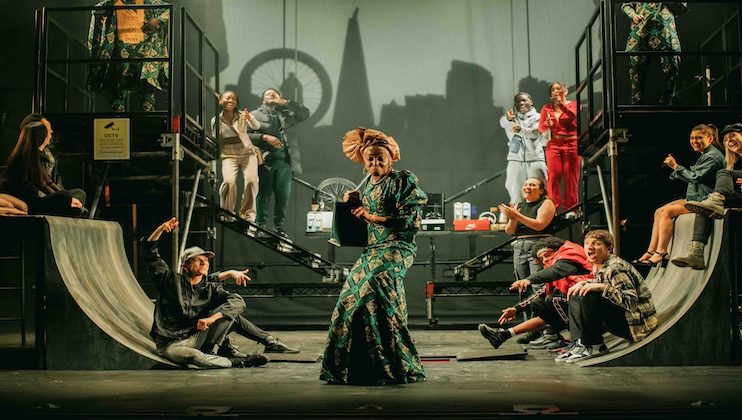 A theater performance with several actors on stage. The central figure is a person in a green patterned costume and headwear, flanked by actors dressed in contemporary clothing on raised platforms and ramps. The backdrop features the skyline silhouettes of iconic buildings.