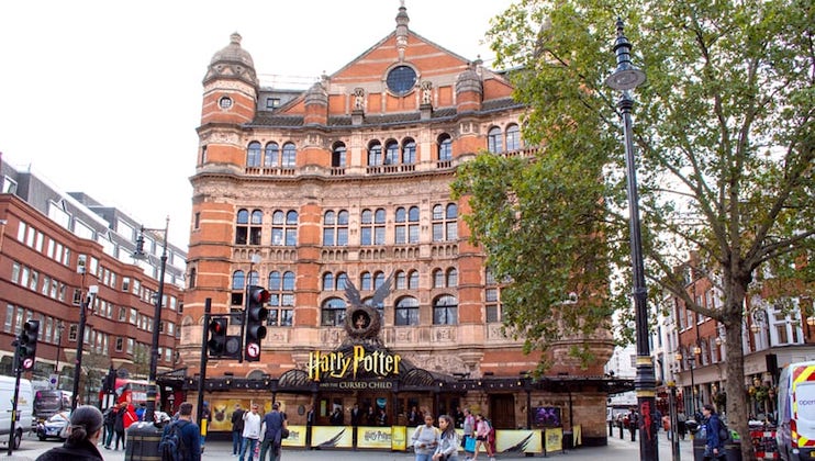 A large, historic theater with an ornate facade and multiple arch windows. The marquee displays Harry Potter and the Cursed Child. People are walking on the street, and a large tree partially obscures the right side of the building.