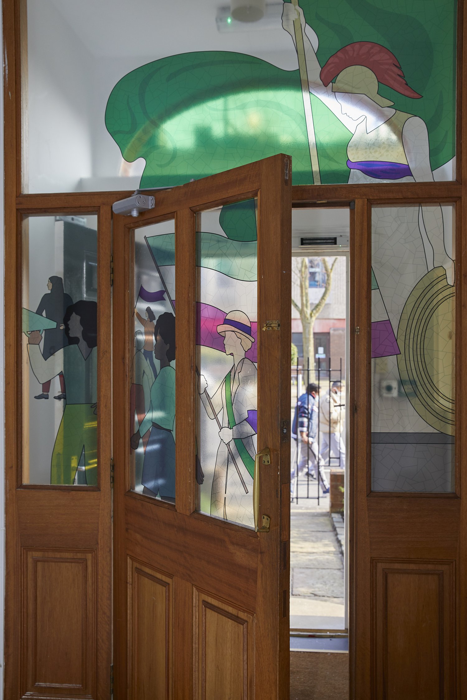 A wooden door with glass panels features colorful stained glass artwork. The designs depict abstract figures, including a woman with a sword and shield, and other people in various poses. Sunlight illuminates the artwork, casting vibrant hues and creating a welcoming atmosphere.