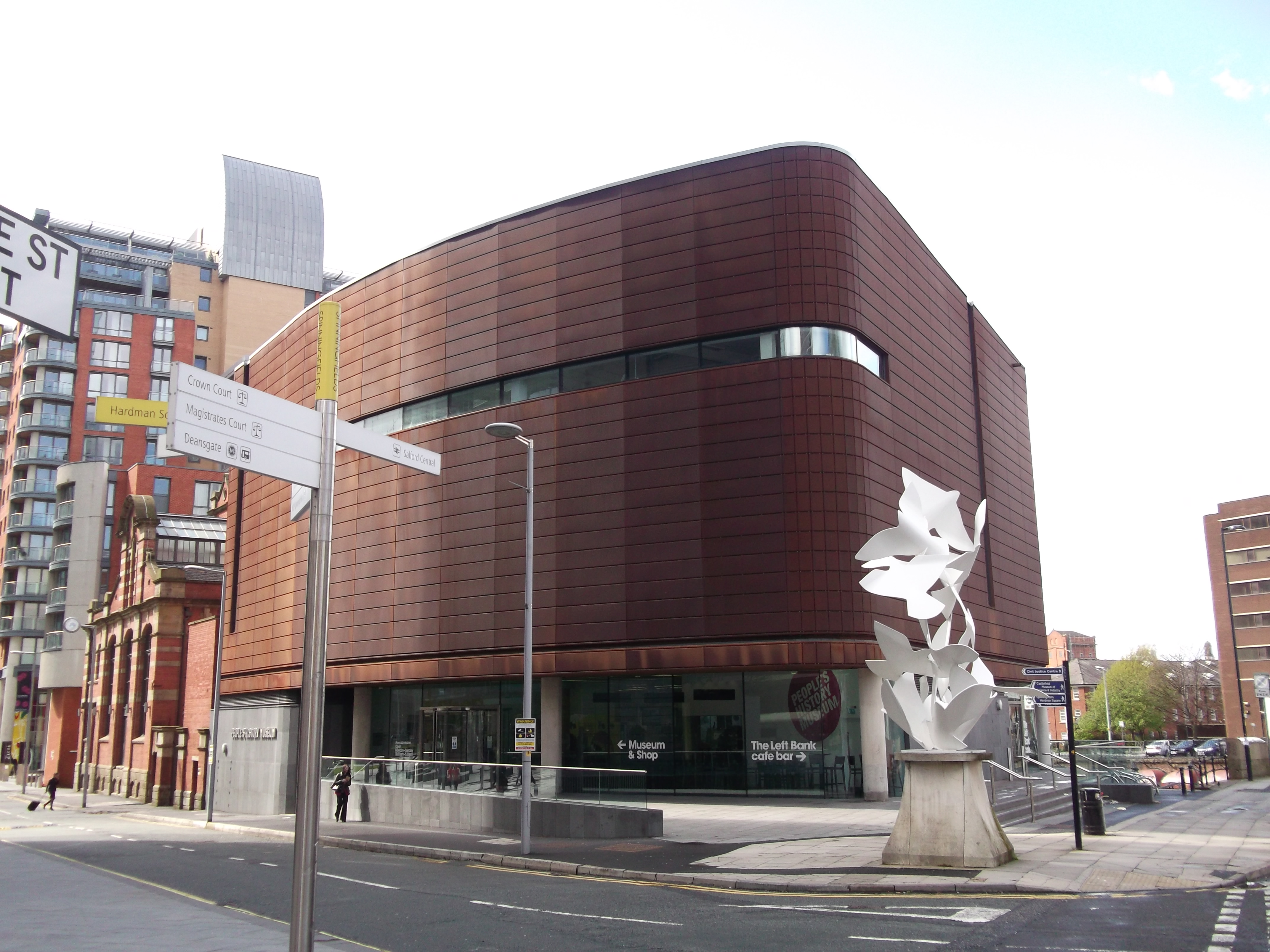 A modern, curved brown building with large windows, housing the People's History Museum in Manchester, UK. There is a white abstract sculpture in front, and street signs indicating nearby streets such as Left Bank. The surrounding area has other urban buildings.