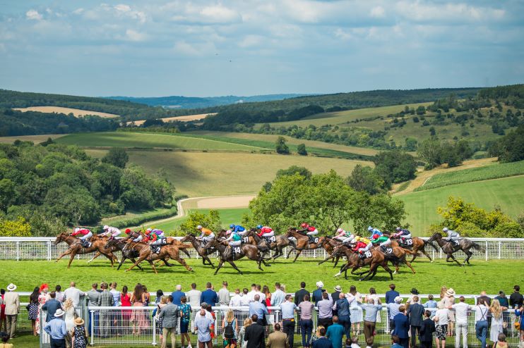 A lively horse race unfolds on a green track set against scenic rolling hills under a blue sky. Jockeys in colorful uniforms ride closely packed horses, while spectators in summer attire watch intently from behind a white fence.