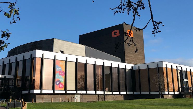 A modern building with floor-to-ceiling windows and brown brick walls. Colorful banners are displayed on the left side of the building. The logo features a stylized letter Q in orange on a dark structure. There is a grassy area with bare tree branches in the foreground.