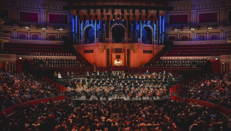 A symphony orchestra performs on stage in an ornate concert hall with an audience seated on the ground level and in balconies. The backdrop features an organ and illuminated architectural details. The hall is filled with people attentively watching the performance.