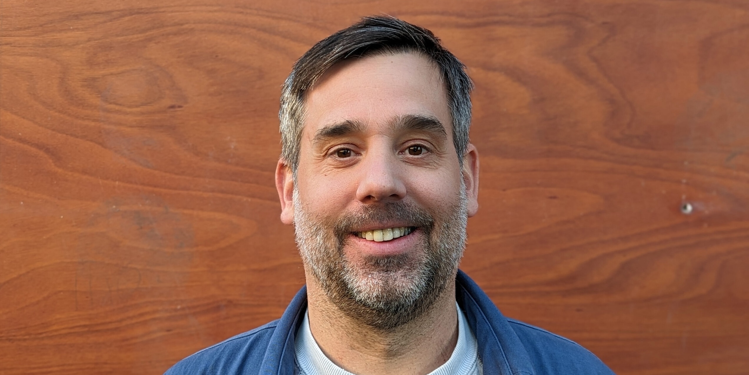 A man with short dark hair and a light beard and mustache is smiling. He is wearing a blue jacket over a white shirt. The background is a wooden surface with natural grain patterns.