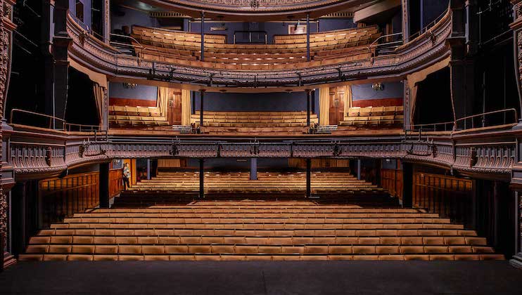 A view from the stage of an empty theater showing rows of seats on the main floor and two balconies. The theater has ornate architectural details with decorative edges and warm lighting, creating an inviting atmosphere. The seats are upholstered in a light color.