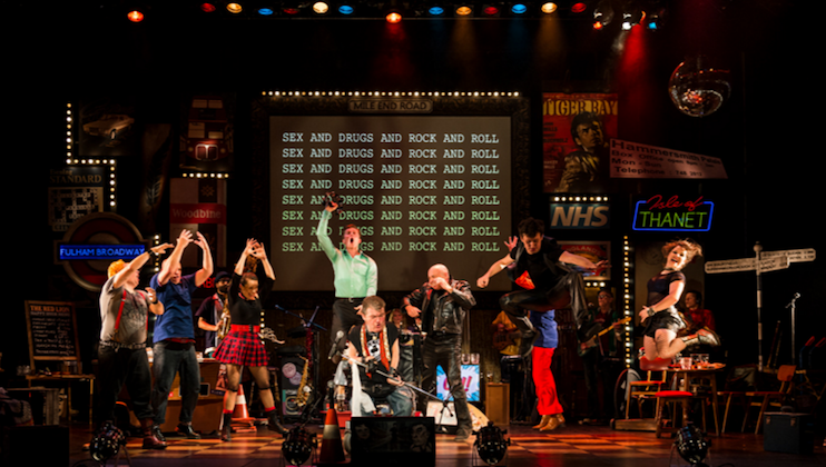 A diverse group of performers on a colorful, illuminated stage. They are energetically posed in various action stances while the background shows a large screen displaying SEX AND DRUGS AND ROCK AND ROLL repeatedly. The stage is filled with vibrant signs and props.