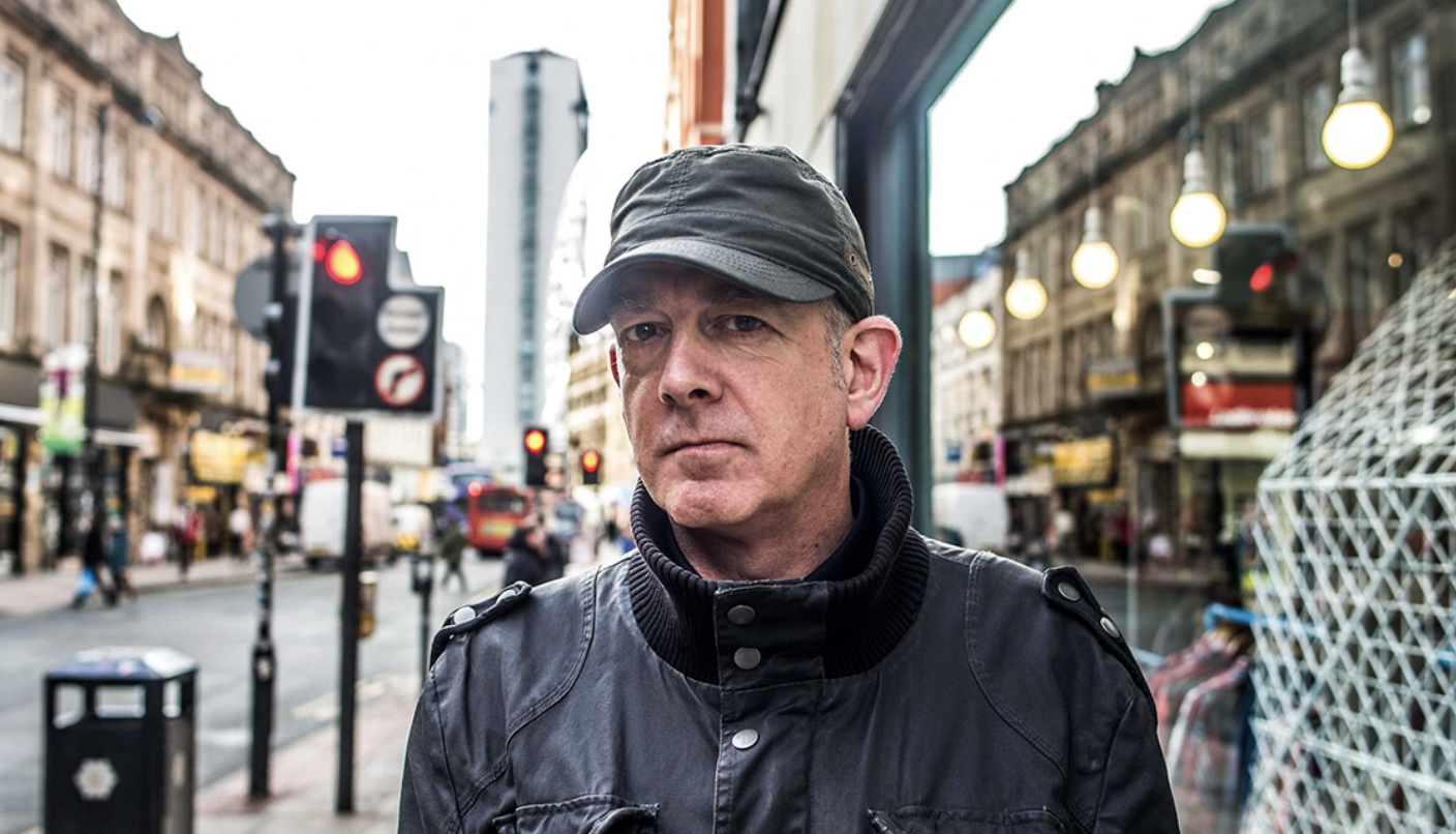 A middle-aged man with a serious expression stands on a bustling city street. He is wearing a grey cap and a dark jacket. Behind him, old buildings line the street, and modern skyscrapers are visible in the background. There are traffic lights and street signs nearby.