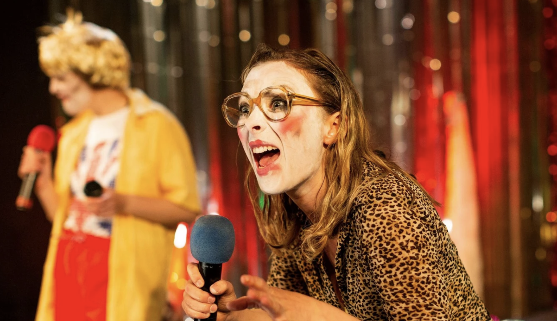 A person is dressed in a leopard-print shirt and large round glasses, holding a blue microphone and wearing clown makeup with white face paint and red cheeks. Another person in the background is wearing a yellow shirt and blonde wig, holding a red microphone.