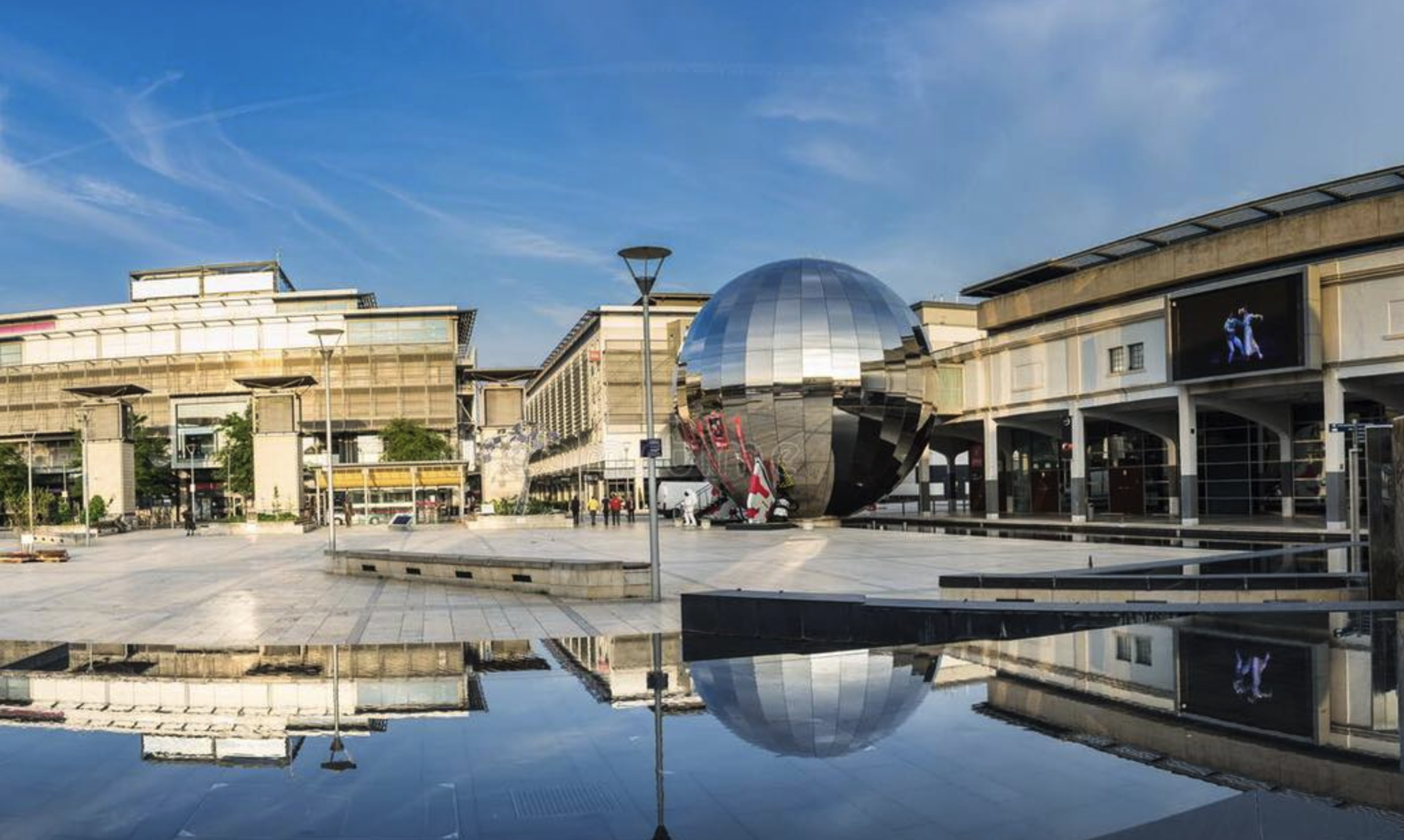 Image of an urban plaza with modern buildings surrounding a large, reflective, spherical structure at its center. The sky is blue with few clouds, and there's a clear reflection of the scene on a shallow body of water in the foreground.