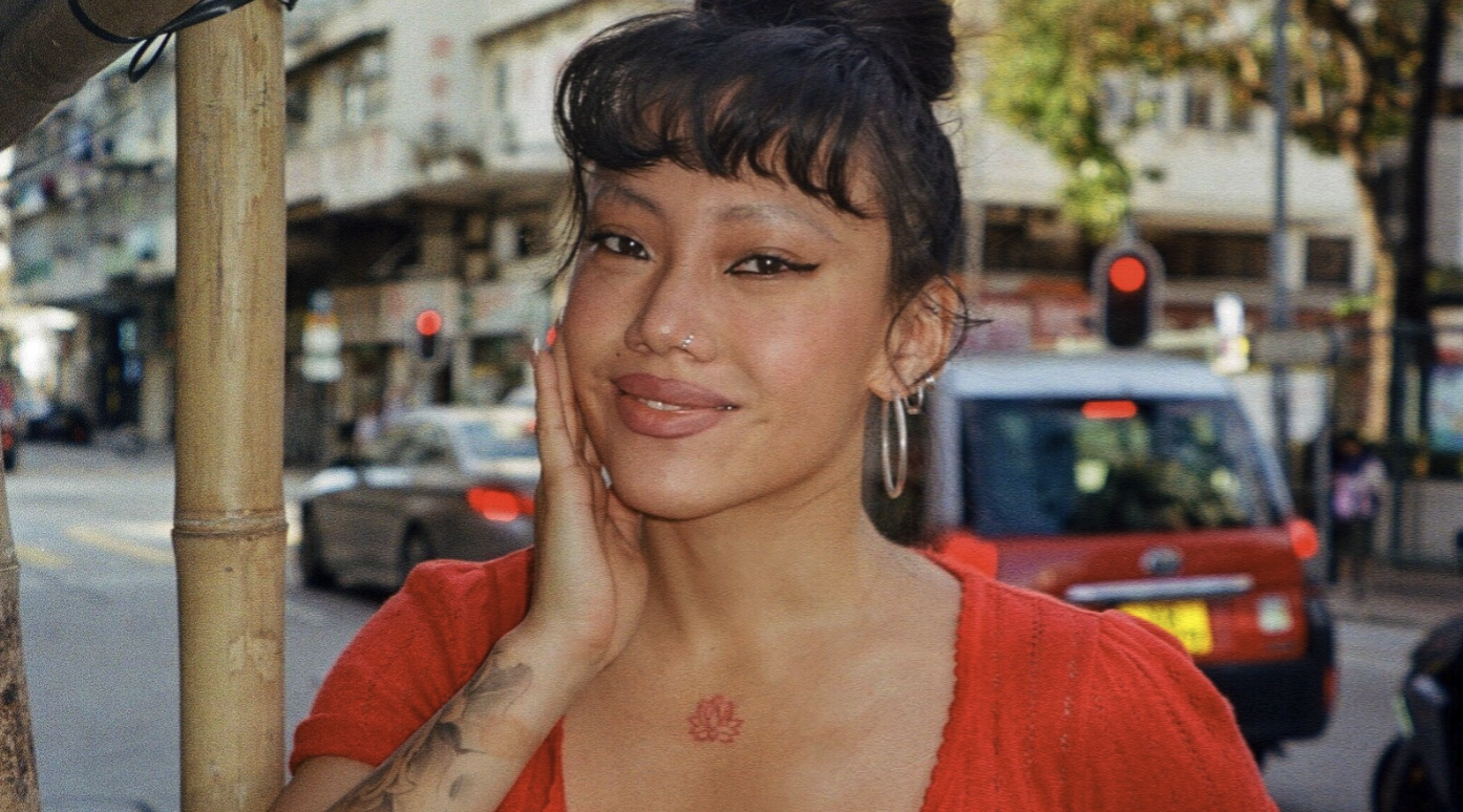 A person with long dark hair styled in an updo, wearing hoop earrings and a red top, smiles while holding one hand to their face. They have visible tattoos on their arm and chest. The background shows a busy street with buildings, cars, and traffic lights.
