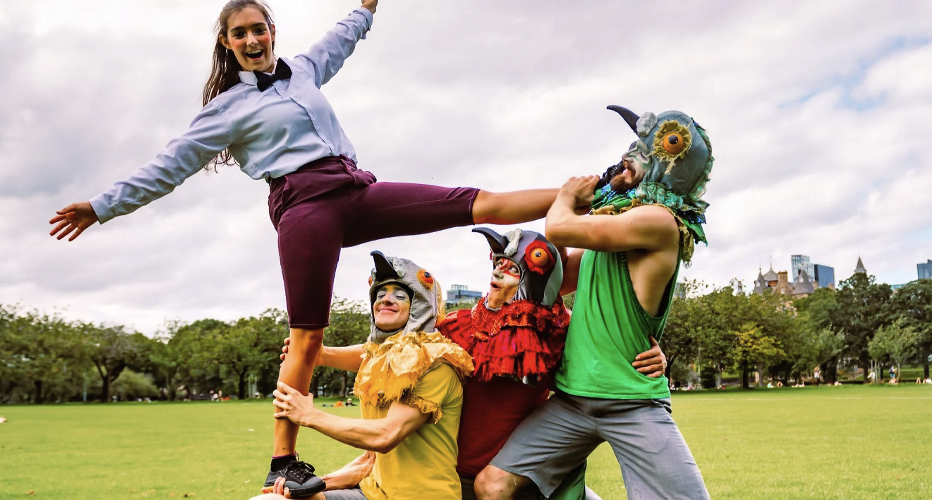 A woman in a blue long-sleeve shirt and purple pants balances on the shoulders of a person in a yellow shirt wearing a bird-like mask. Two others, also wearing bird masks, in green and red shirts, support her. They are outdoors on a grassy field with trees in the background.