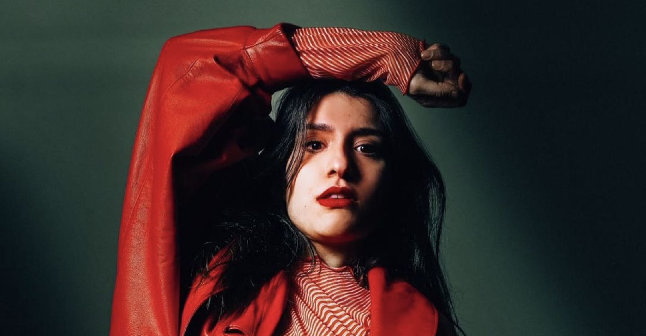 A person with long dark hair, wearing a red jacket over a striped top, poses with their left arm raised and bent at the elbow, casting a shadow. The background is dark and the lighting creates a dramatic effect on their face.