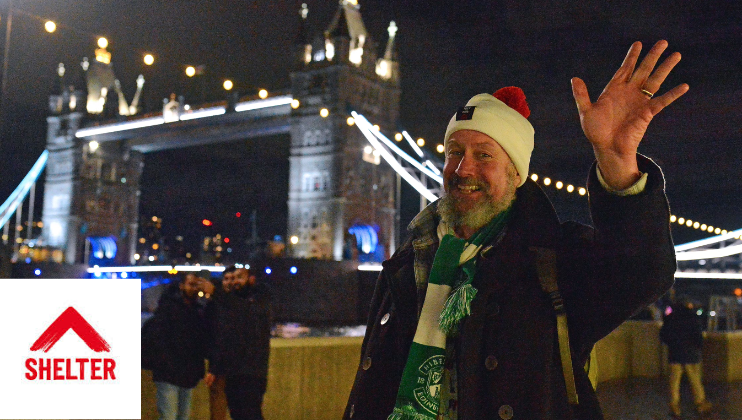 A man in a Santa hat waves to the camera; he is walking the Walk for Home for the homelessness charity Shelter - in the background is Tower Bridge, lit up against the night sky. In the bottom left of the image is a small Shelter UK logo