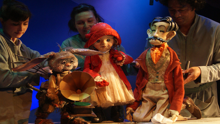 Three puppeteers manipulate and present four puppets on a stage. The puppets include a rabbit with a gramophone, a doll in a red bonnet and dress, a gentleman with a mustache and elegant clothes, and a small creature with elongated ears. The background is blue.