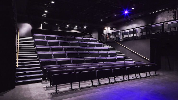 A modern, empty theater with a raked seating arrangement featuring rows of black cushioned seats. The theater has dark walls, ceiling, and flooring, with a well-lit stage area and an upper balcony accessible by stairs. The overall ambiance is sleek and contemporary.