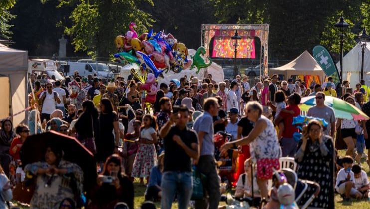 A large crowd gathers outdoors at a summer festival, with people sitting on the grass and walking around. Various stalls, tents, and a first aid station are visible. Colorful balloons are being held in the air, and trees can be seen in the background.