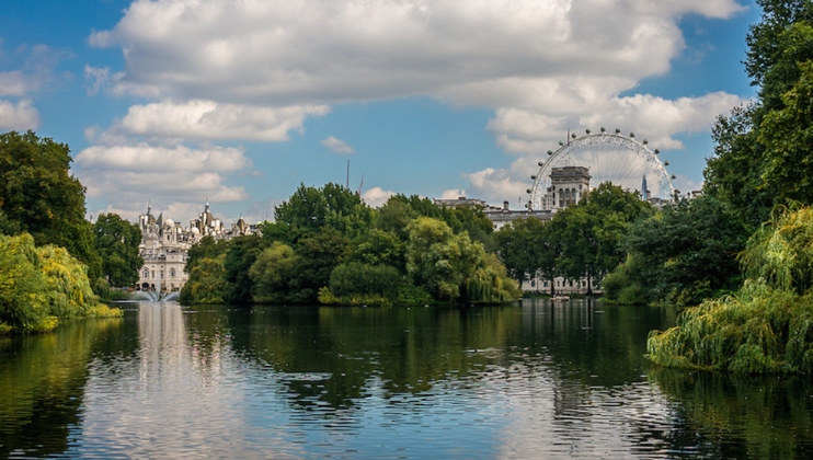 A serene park scene with a calm lake surrounded by lush green trees under a blue sky with scattered clouds. A large Ferris wheel and historic buildings are visible in the background, reflecting in the water.