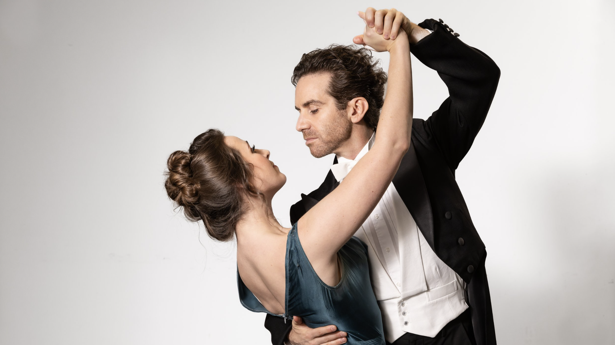 A man and a woman in formal attire are dancing gracefully. The man, in a black tuxedo with a white bow tie, holds the woman's hand above his head while gazing into her eyes. The woman wears an elegant teal dress, her hair styled in an updo. They stand against a neutral white background.