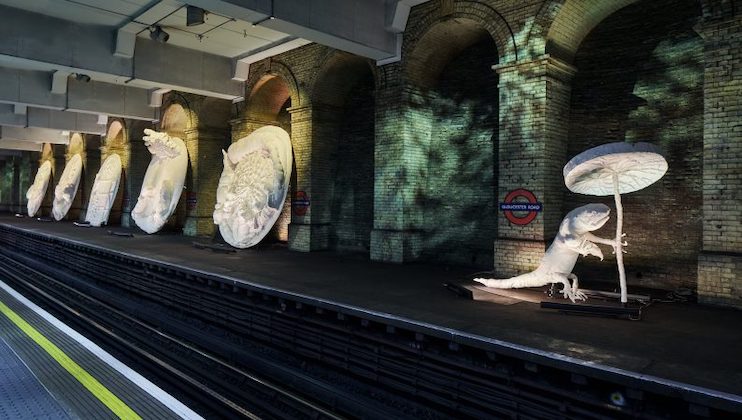 A subway platform at Gloucester Road station in London displays large white sculptures of lizards and various shapes under spotlights. The sculptures cast dramatic shadows against the brick archways. The station name is visible in the background on red and blue signs.