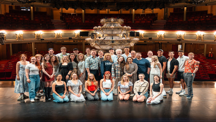 A group of people stands and kneels on a theater stage, smiling for a photo. They form multiple rows with a chandelier prop in the background. The theater seats and ornate balcony railings can be seen behind them.