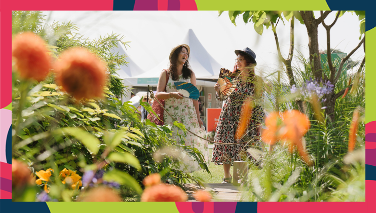 Two women happily walk through a lush garden filled with vibrant flowers and plants. One woman wears a patterned dress and hat while the other wears a floral dress and sunhat. Both are smiling and engaging in conversation. A white tent is visible in the background.