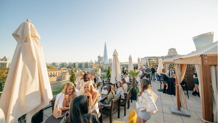 People are gathered on a rooftop bar during a sunny day, enjoying drinks and conversation. The view includes a cityscape with tall buildings in the background. Tables with umbrellas and greenery are scattered around the terrace, creating a lively yet relaxed atmosphere.