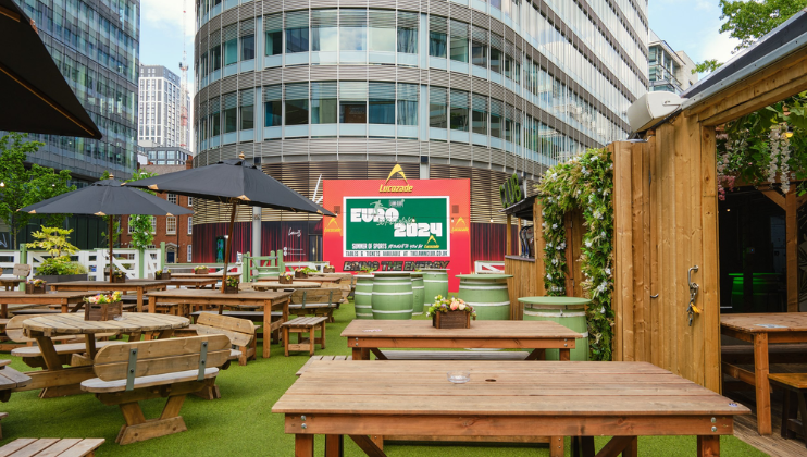 An outdoor seating area with wooden picnic tables and benches on artificial grass is shown. Large umbrellas provide shade. In the background, a big screen displays the Euro 2021 event. Buildings and greenery surround the space, giving it an urban yet relaxed feel.