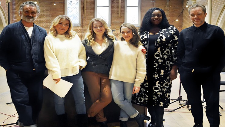 A group of six people standing in a row, posing for a photo inside a room with high ceilings and arched windows. They are dressed in casual to semi-formal attire, smiling at the camera. There are microphones and stands visible in the background.