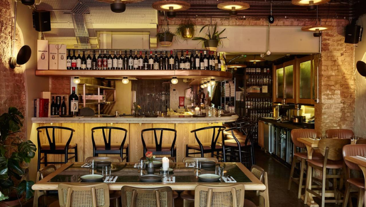 A cozy restaurant interior featuring a wooden bar lined with bottles of wine, black chairs, and industrial-style lighting. The dining area includes a table set for four with placemats and plates. Exposed brick walls and lush plants add a warm ambiance.