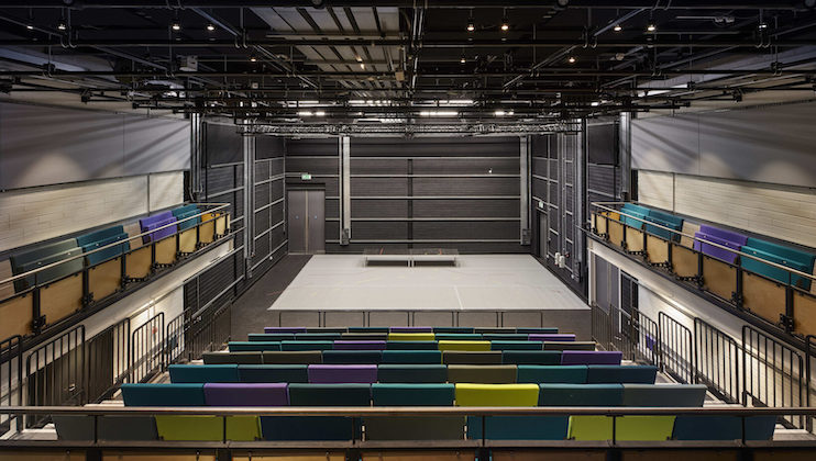 The image shows an interior view of an empty, modern theater with tiered seating. The seats are a mix of purple, green, and blue colors. The stage is simple, with a gray floor and a centered rectangular platform. Overhead lighting and a backstage door are visible.