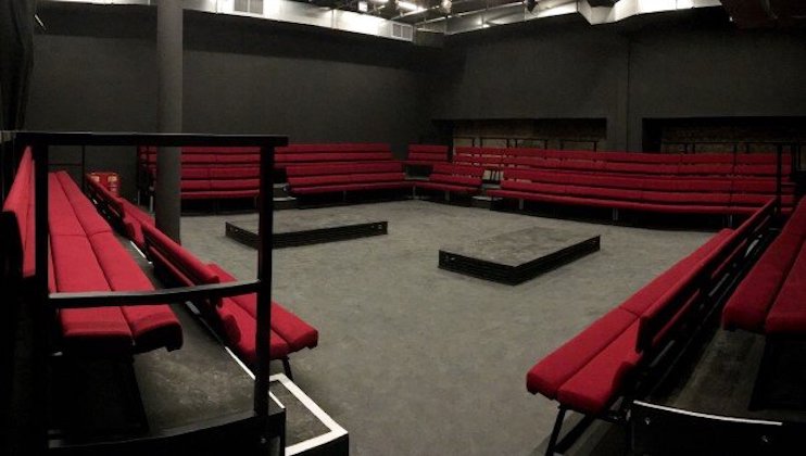 A black box theater is shown with red seating arranged in a square around an empty central performance space. The seats are tiered and surrounding the stage on all four sides in a minimalist design. The walls and floor are black, enhancing the dramatic atmosphere.