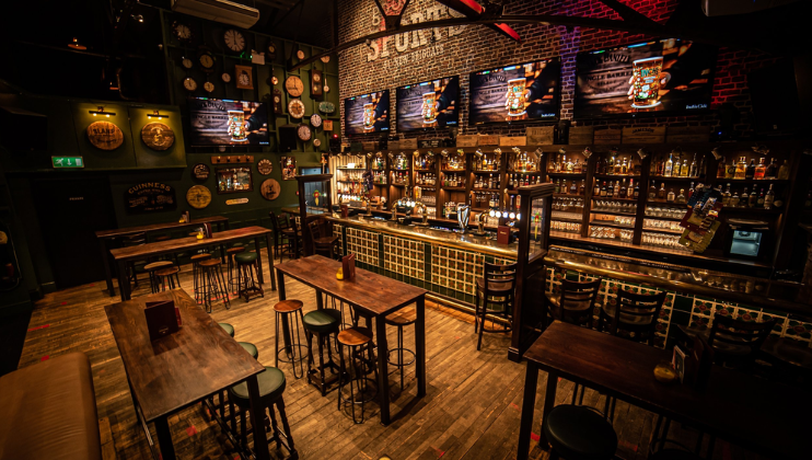 A dimly lit bar with a warm, inviting atmosphere. High wooden tables and stools are scattered throughout. The bar counter in the background is well-stocked with various bottles, and three large TV screens above display a sports game. Decor includes clocks and beer signs.