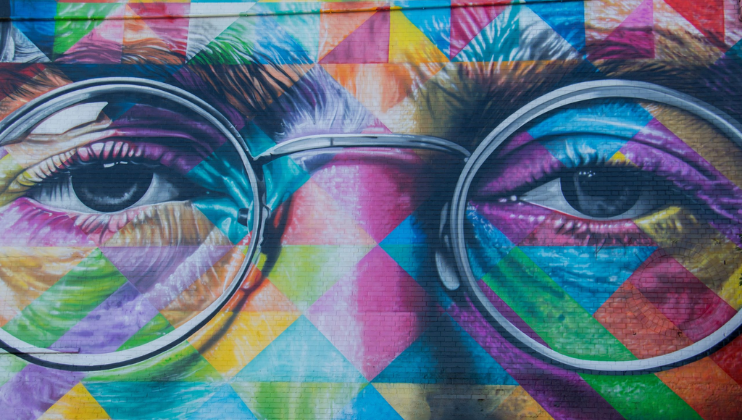 A vivid and colorful mural of a person's face wearing round glasses. The artist uses geometric patterns and a rainbow palette to create a striking, modern design that covers the entire wall. The eyes are particularly detailed, contrasting with the abstract background.