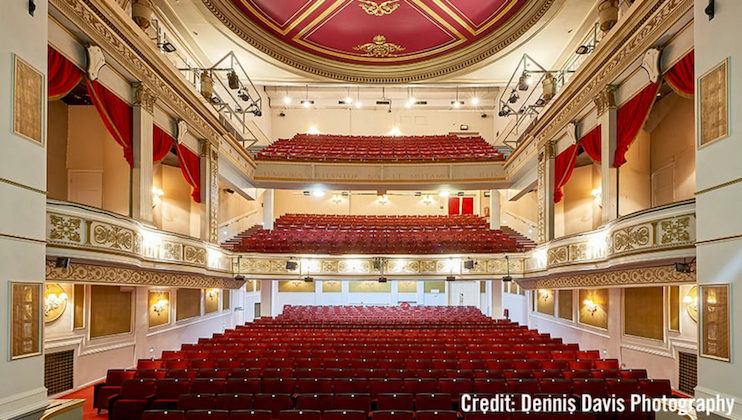 A luxurious, empty theater with red and gold decor. Rows of red seats extend from the stage to the rear balcony. The ceiling features ornate decorations and red curtains adorn the boxes on either side. Photographer credit is given to Dennis Davis.