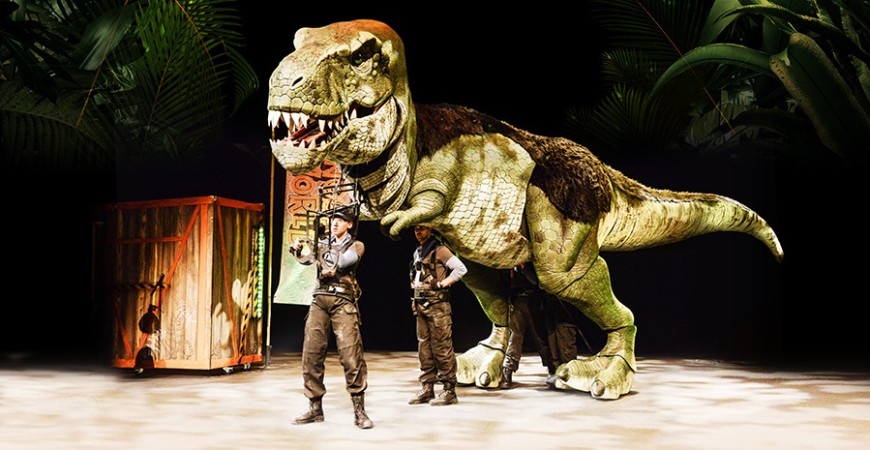 Two performers in adventurer outfits guide a large, realistic Tyrannosaurus rex puppet on stage. Behind them, there is a colorful crate. The scene is set against a backdrop of lush, green jungle foliage.